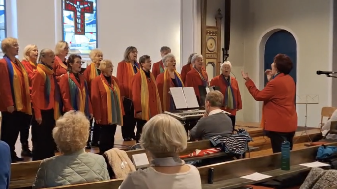 Chor mit bunten Schals singt in einer Kirche, dirigiert von einer Frau in rotem Anzug.